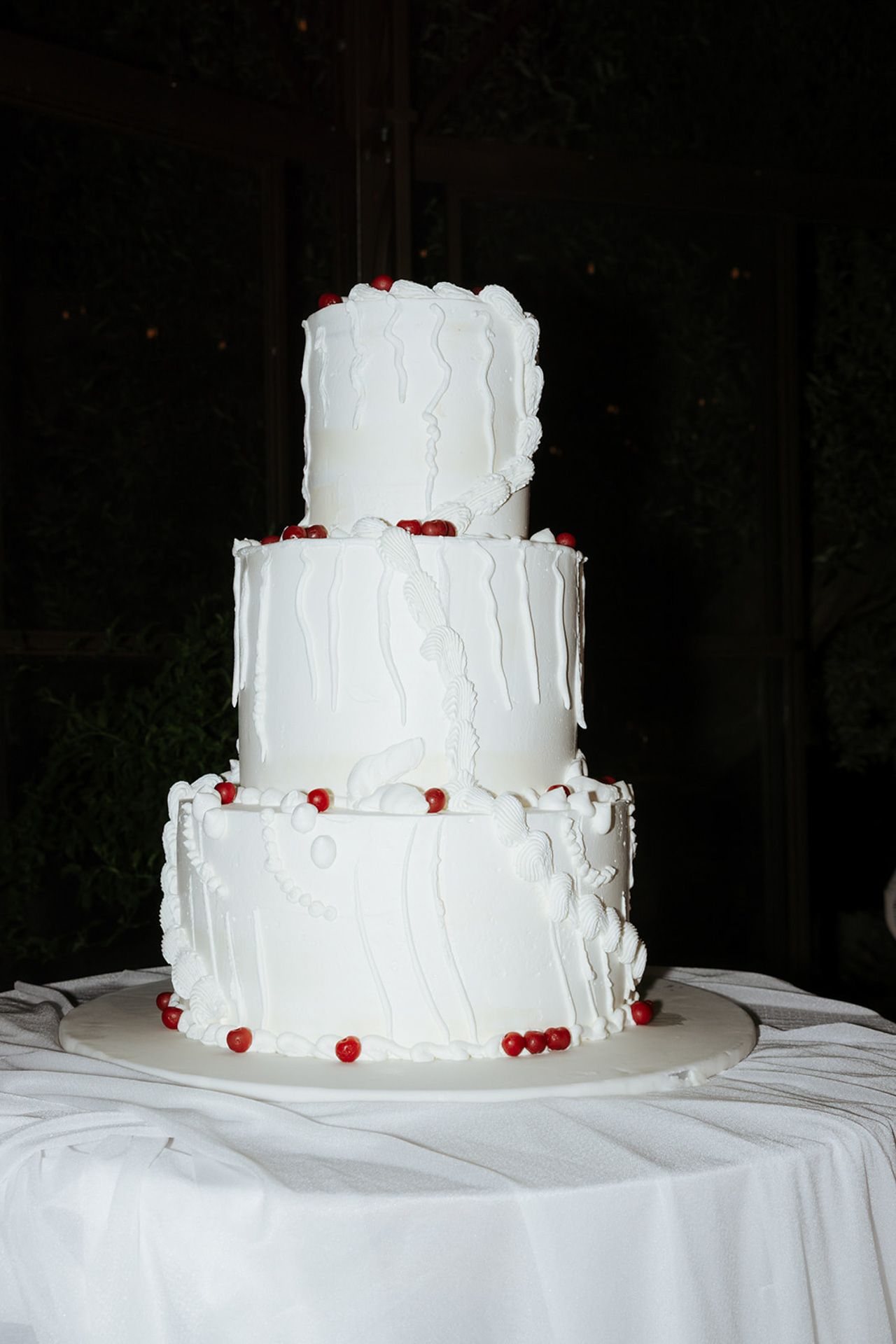 A three-tiered white wedding cake with ruffled frosting and red accents sits on a white tablecloth.