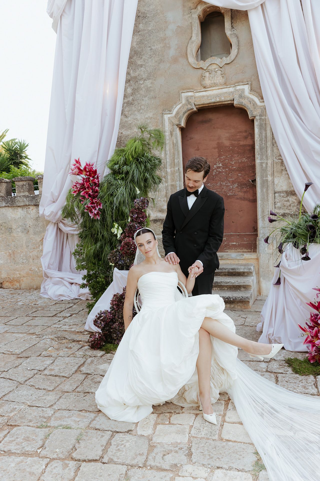 A romantic wedding photoshoot in an elegant stone archway with flowing white curtains and pink floral decorations.