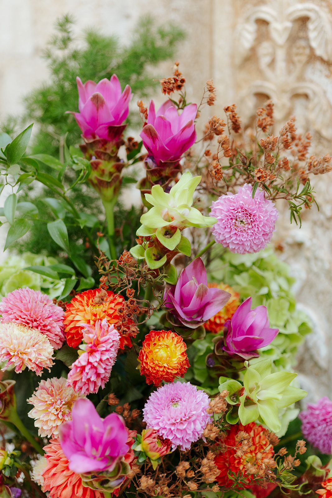 Vibrant arrangement of pink chrysanthemums and bright orange dahlias with green foliage against a beige wall.