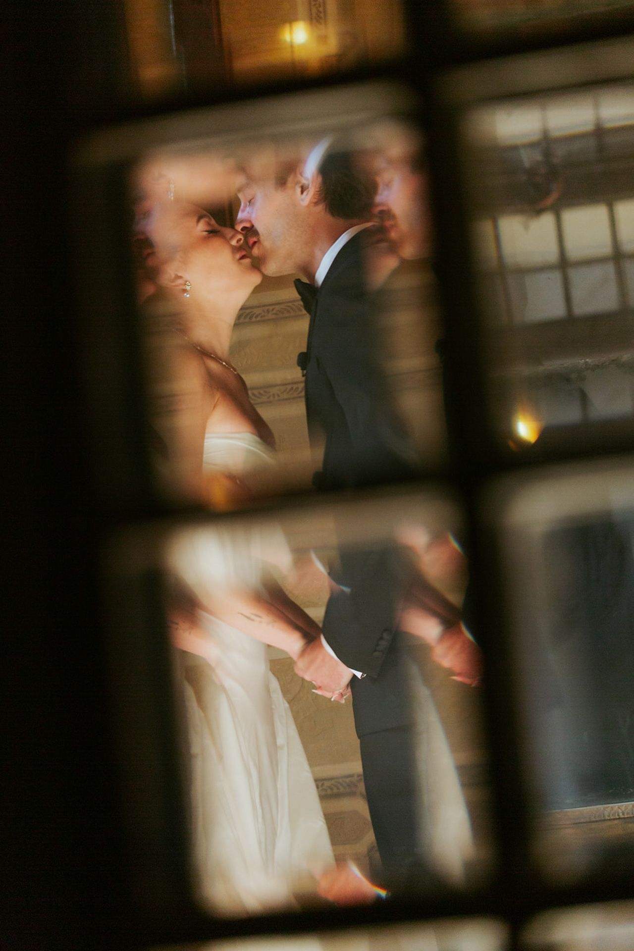 Romantic wedding moment captured through window panes showing couple in white dress and dark suit sharing an intimate kiss.