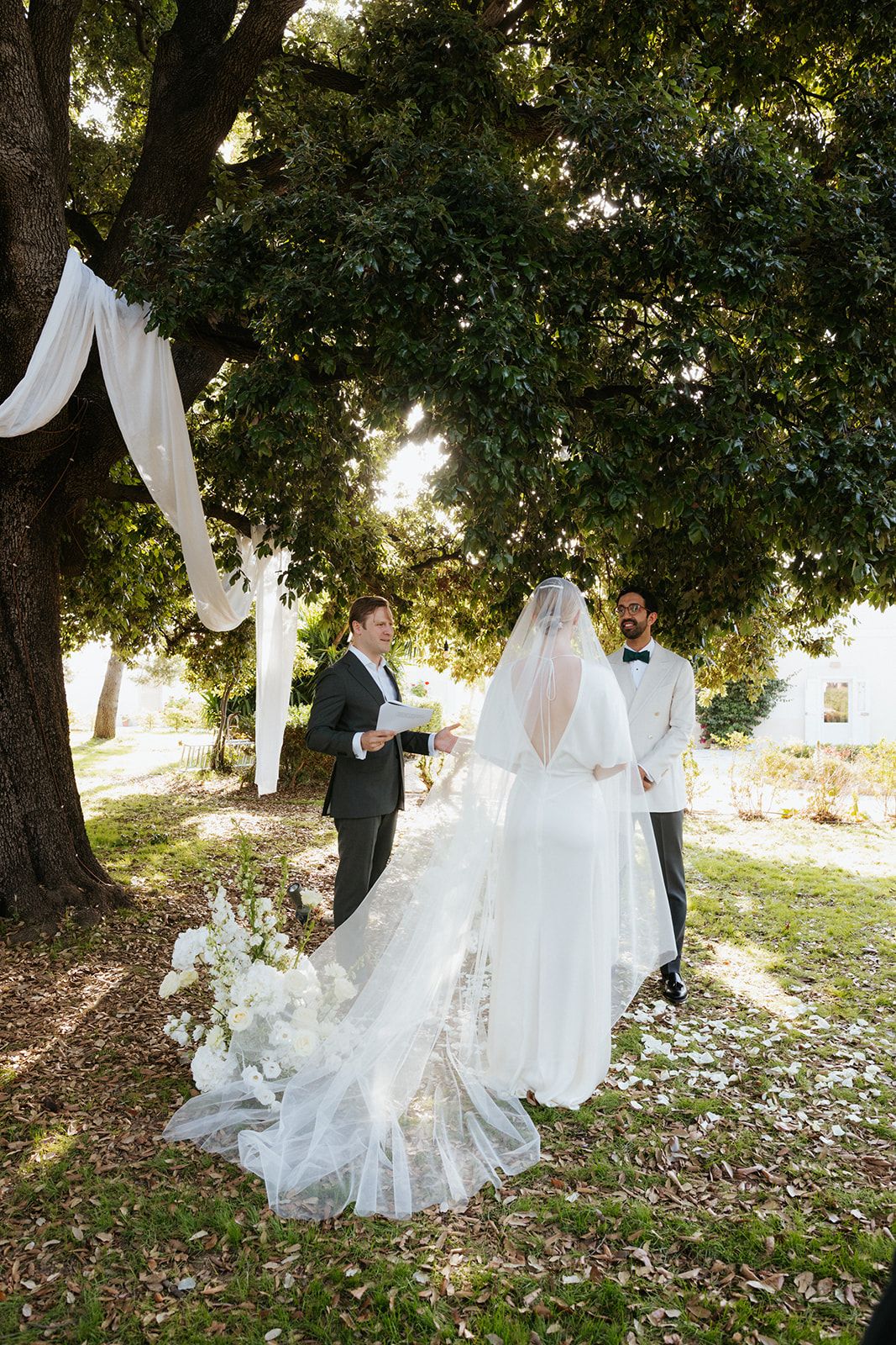 A couple poses together in a dreamy outdoor garden setting with flowing white fabrics and natural greenery.