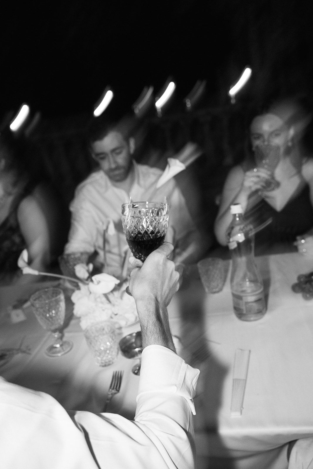 A black and white photograph of raised wine glasses at an elegant dinner party setting.