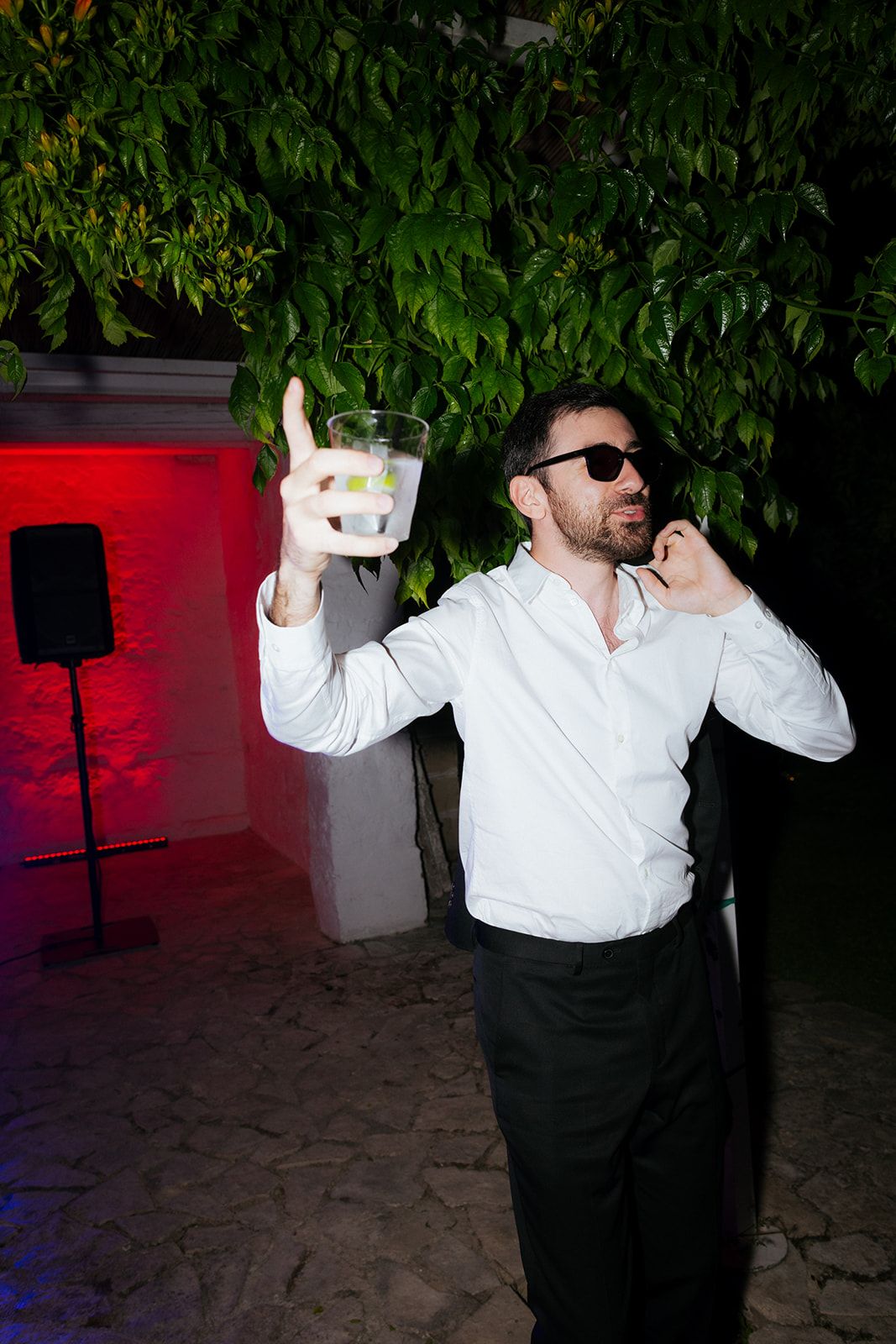 Someone in a white dress shirt and sunglasses holds up a glowing cocktail at a nighttime party with red lighting.