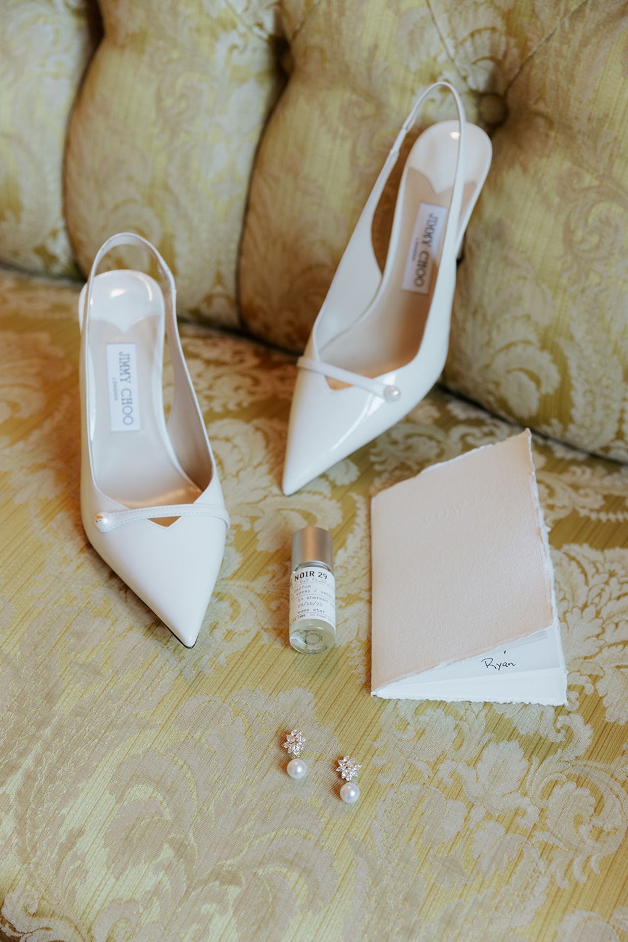 Elegant white pointed-toe bridal shoes and pearl earrings arranged on a patterned beige carpet.