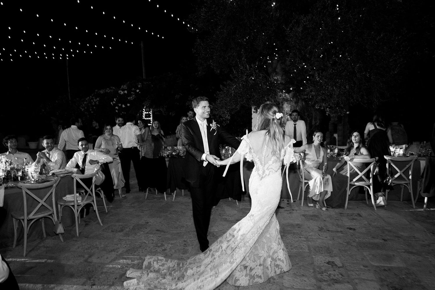 Elegant outdoor evening wedding reception with glowing string lights illuminating guests seated at tables in a garden setting.