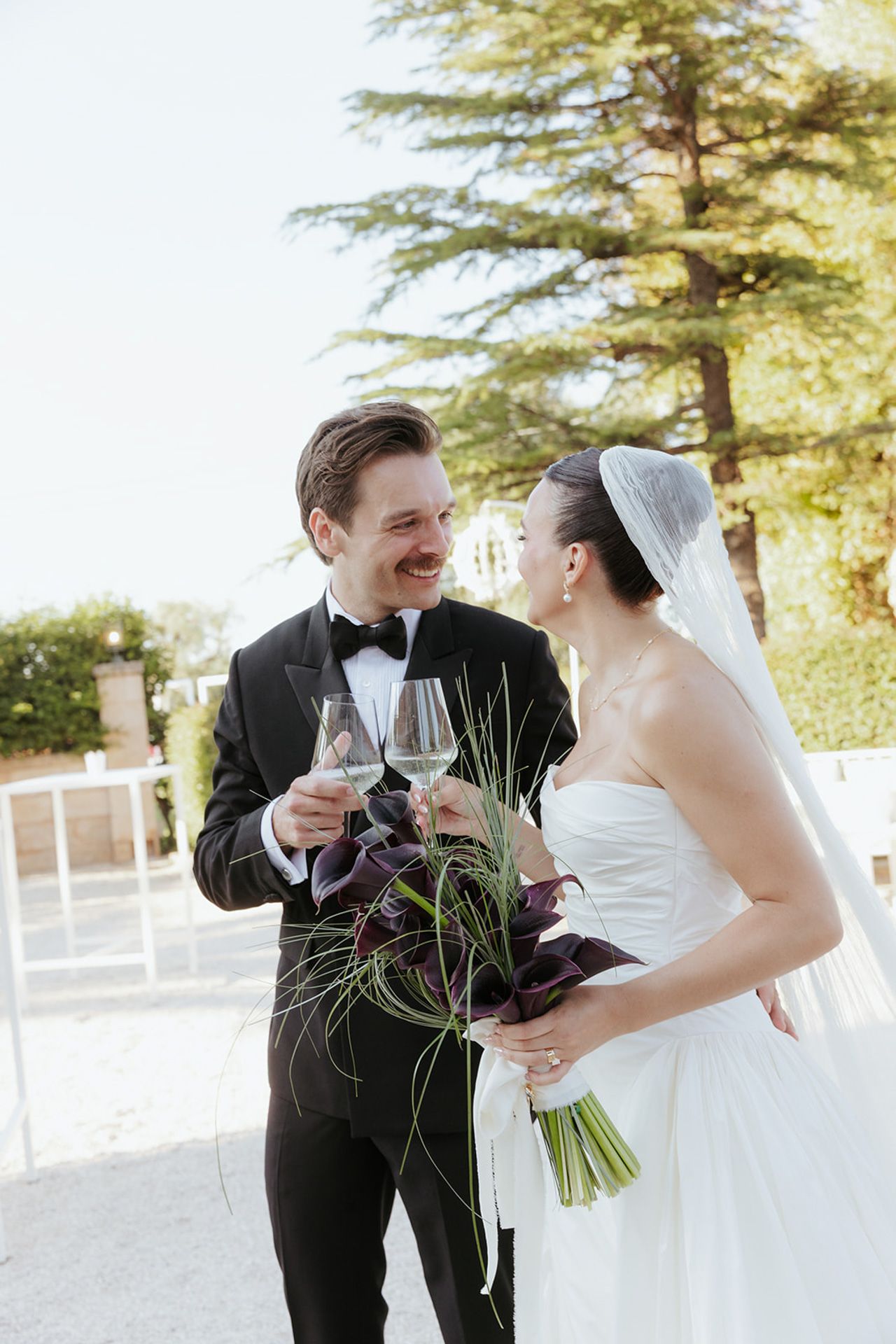A couple shares a joyful moment toasting with wine glasses during their outdoor wedding celebration.