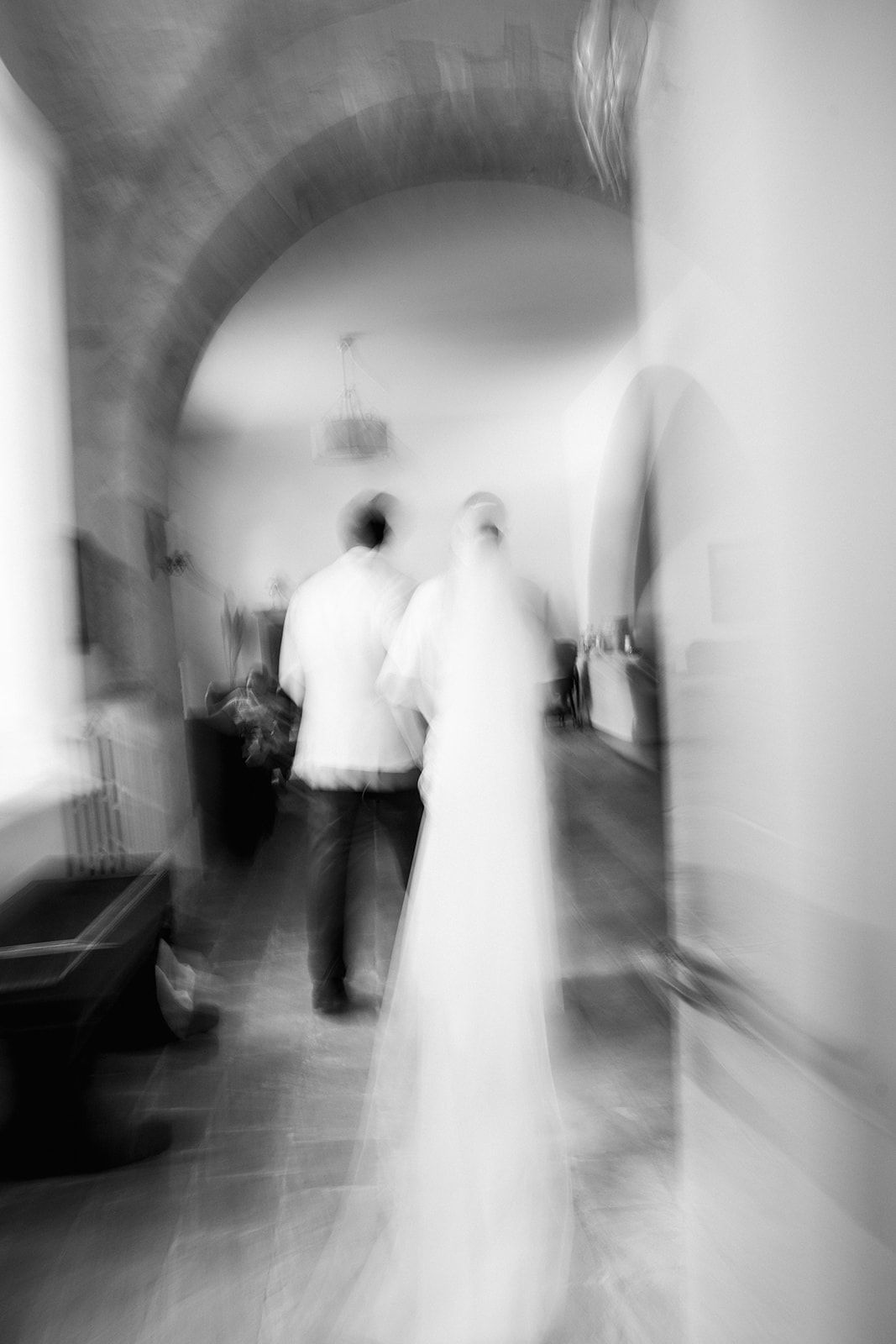 Blurred black and white artistic photograph of a couple in formal attire walking through an arched hallway.