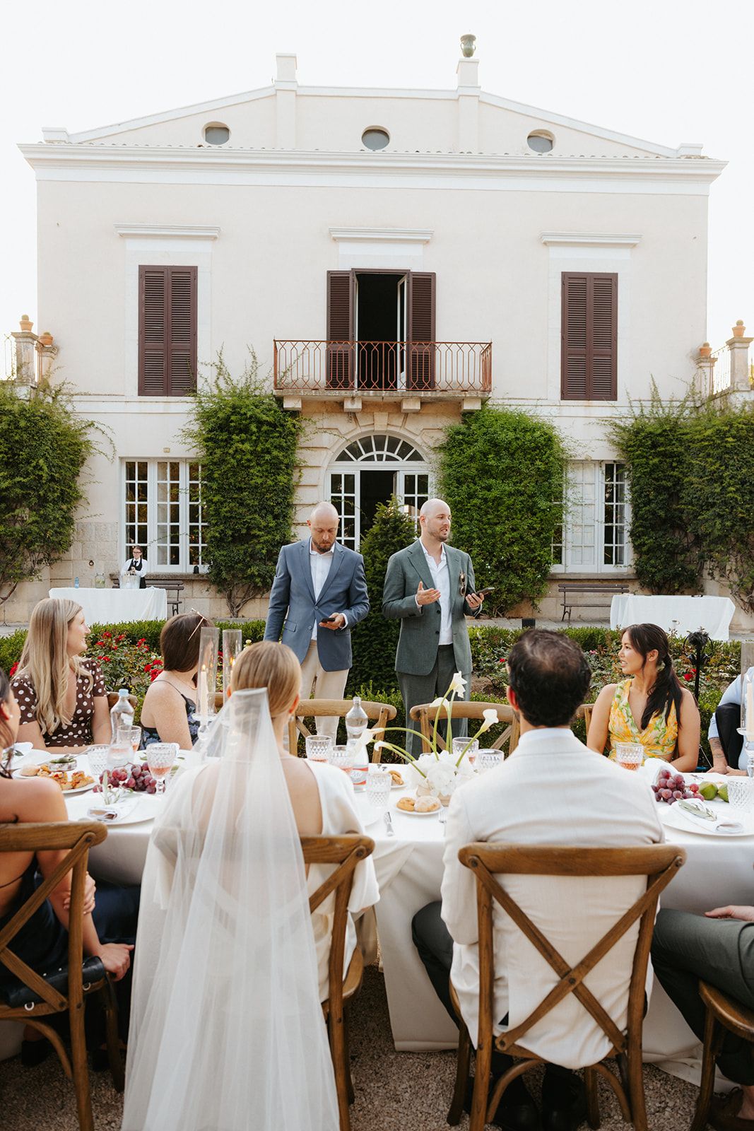 An elegant outdoor wedding reception setup with white tablecloths and farmhouse chairs against a white Mediterranean villa.