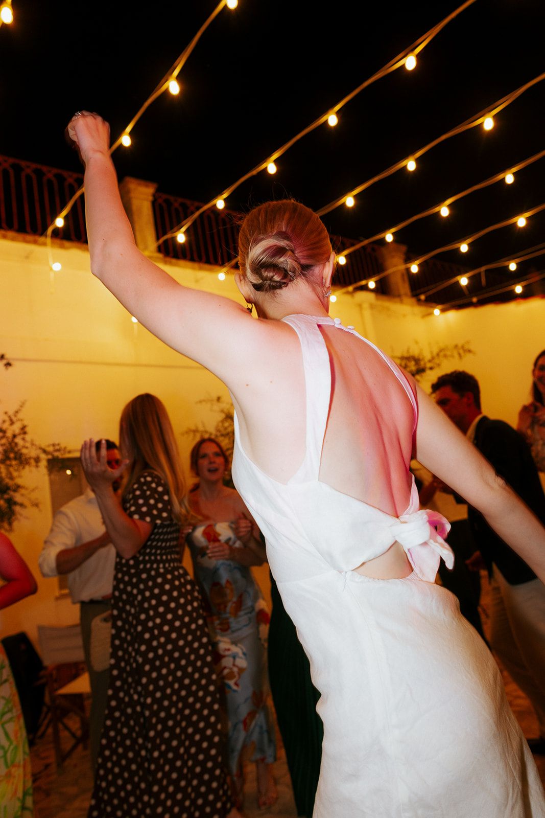 String lights illuminate dancers at a lively party with a person in white dress raising their arms.