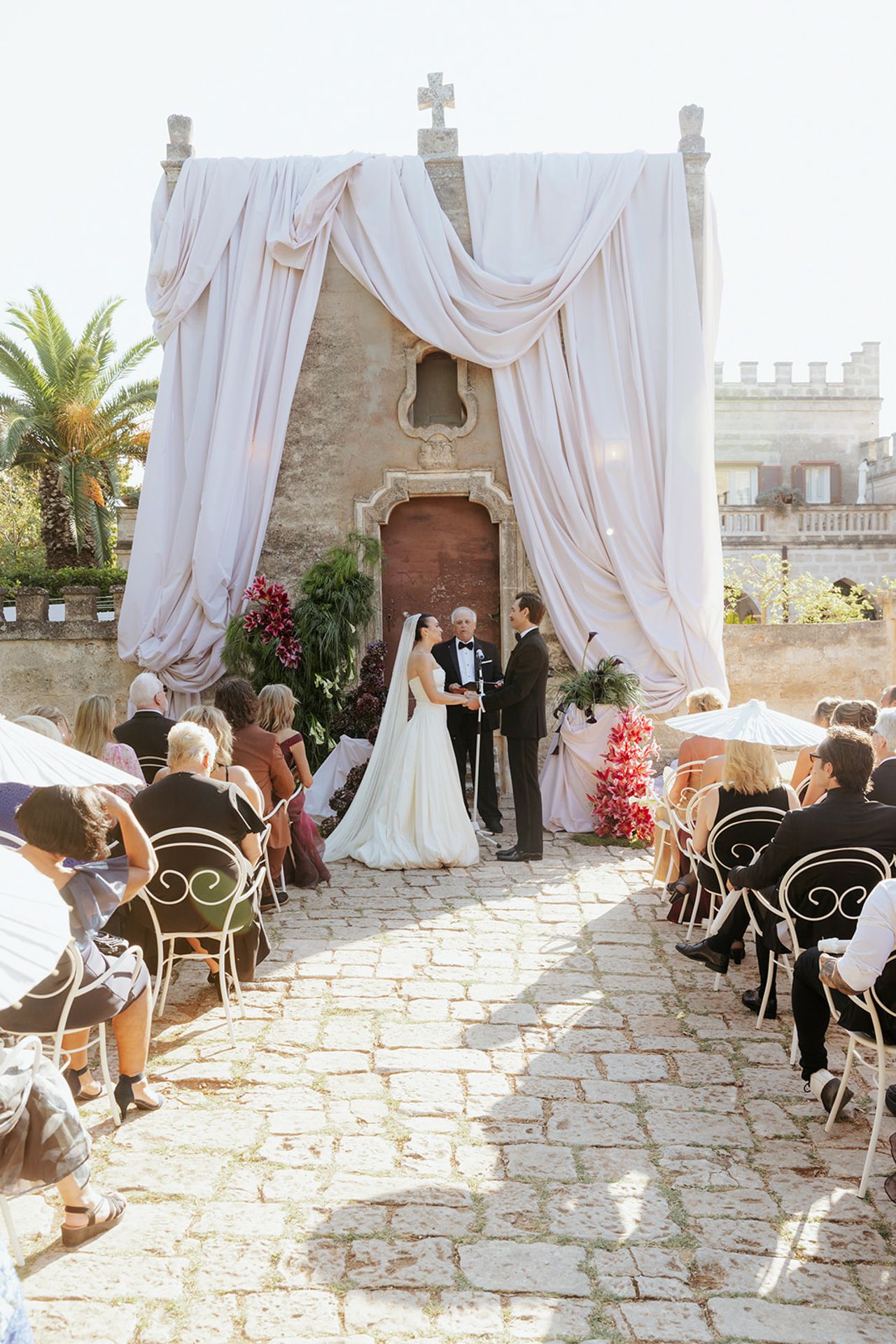 An outdoor wedding ceremony with white draped fabric and elegant black chairs arranged on a stone patio.