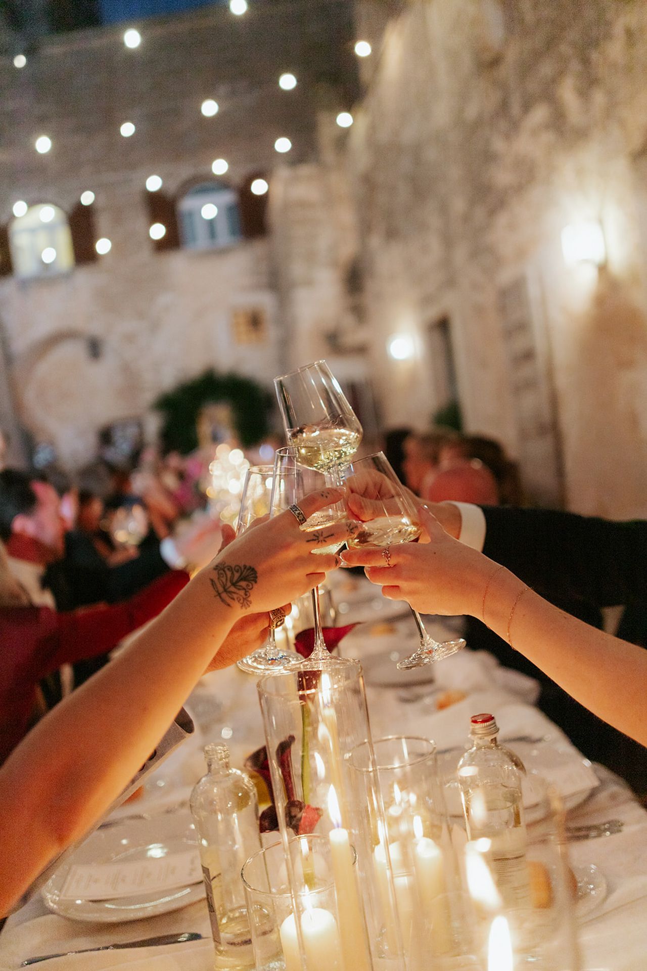 Hands raise wine glasses in a toast across an elegant dinner table with twinkling lights in the background.