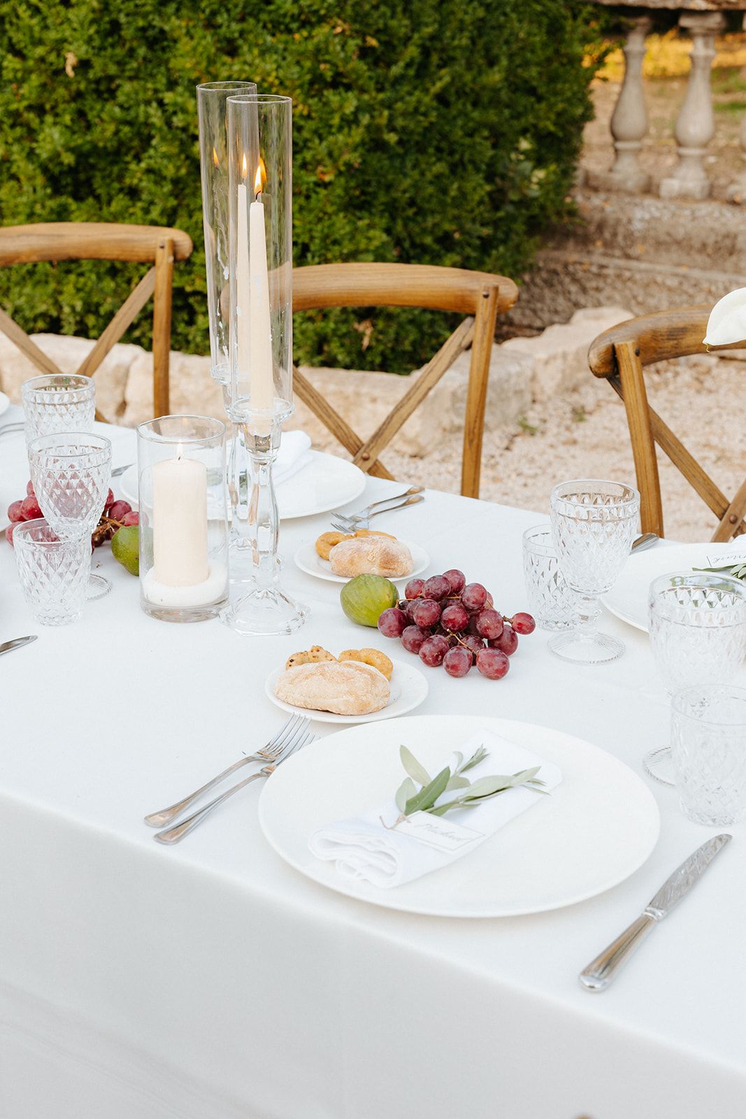 Elegant outdoor table setting with wooden cross-back chairs, crystal glassware, grapes and white linens in garden setting.