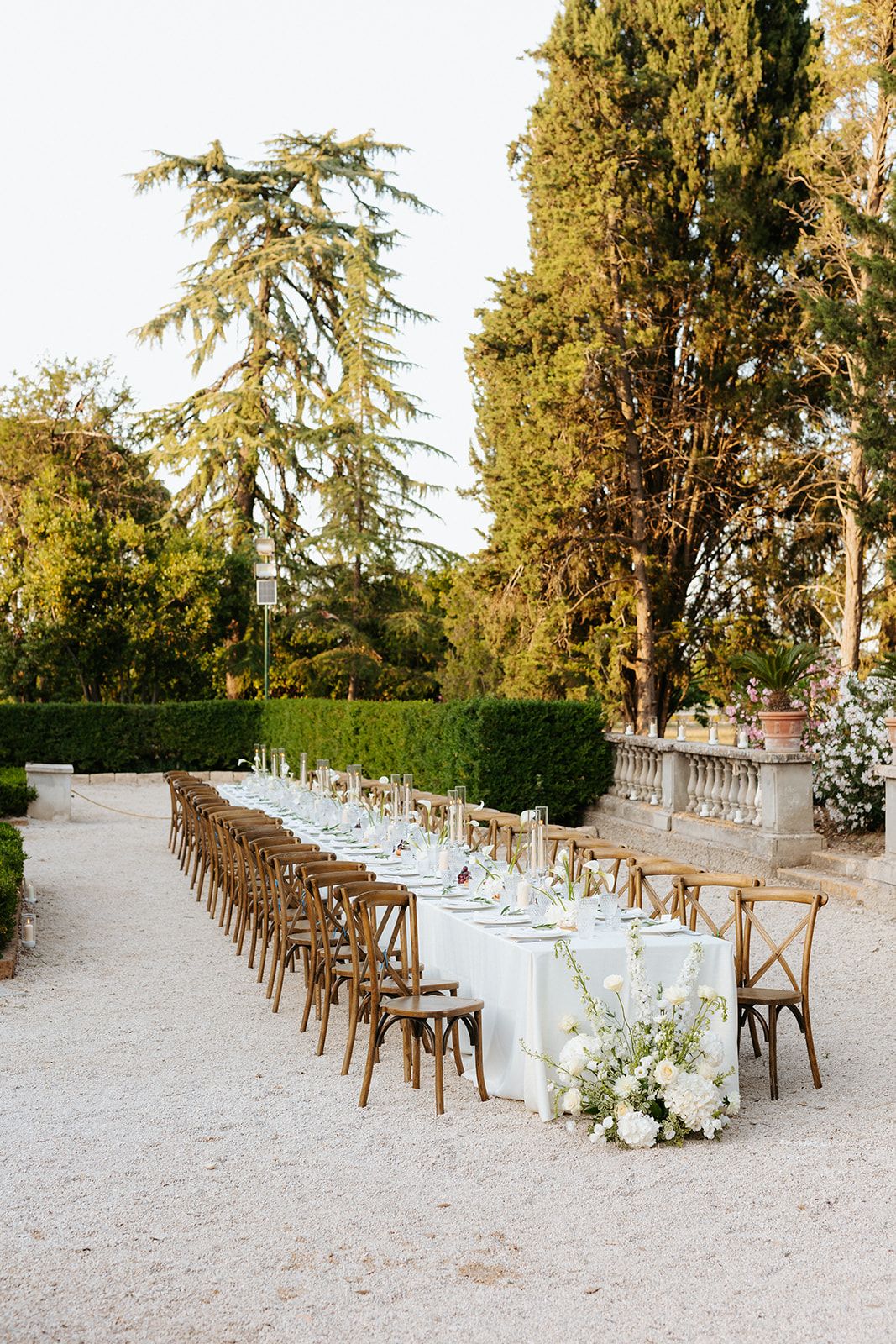 Long white banquet table with wooden chairs set up outdoors along stone balustrade and cypress trees in garden setting.