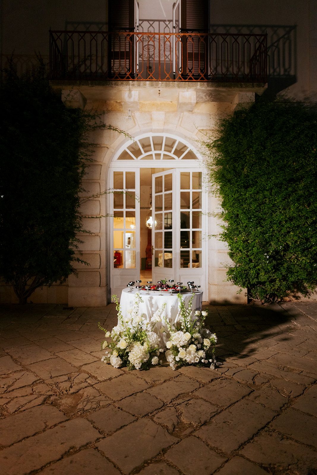 A softly lit stone archway entrance adorned with glowing white floral arrangements and string lights creates an elegant nighttime scene.