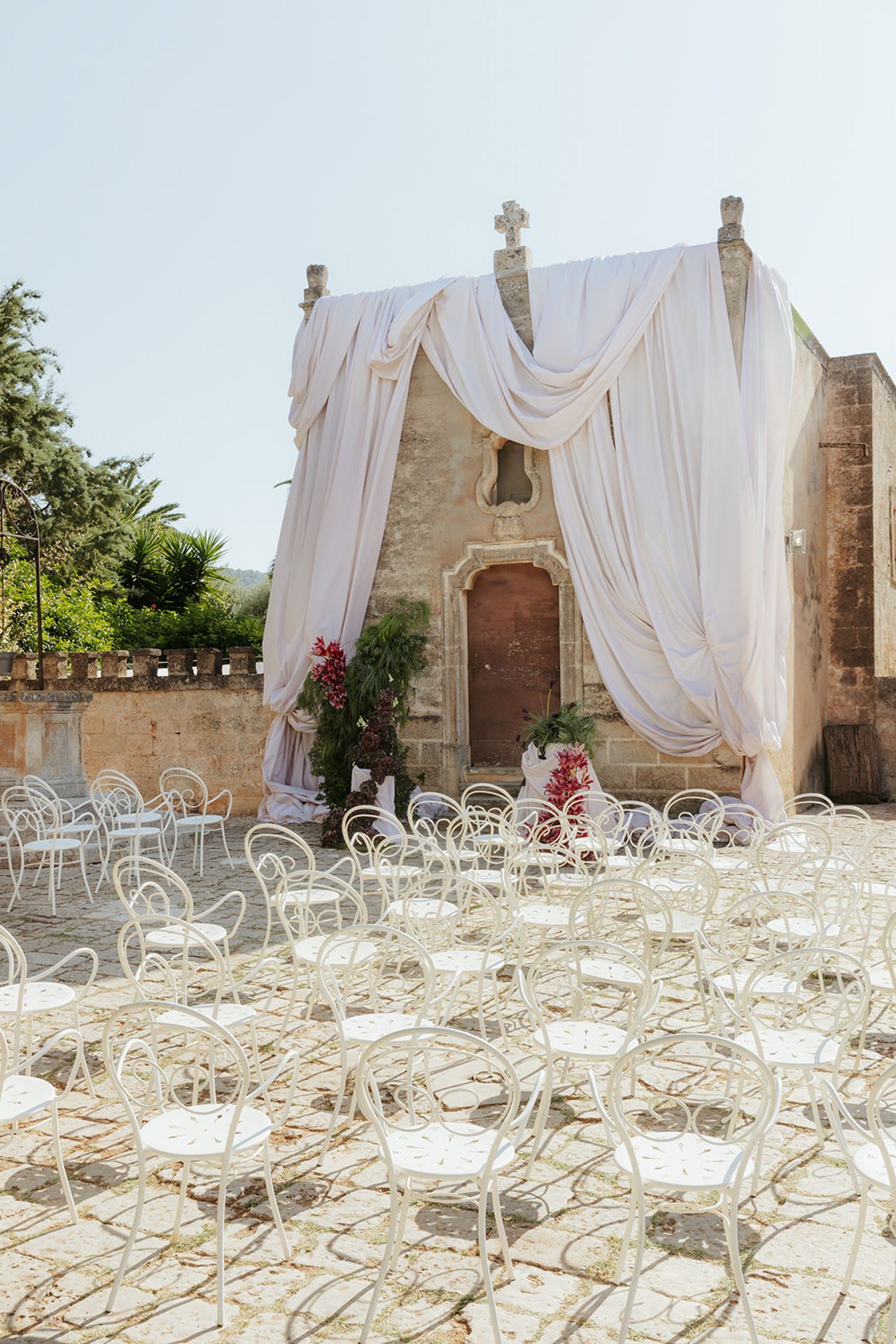 White chairs and flowing fabric drapes create an elegant outdoor wedding ceremony setup against a rustic stone building.
