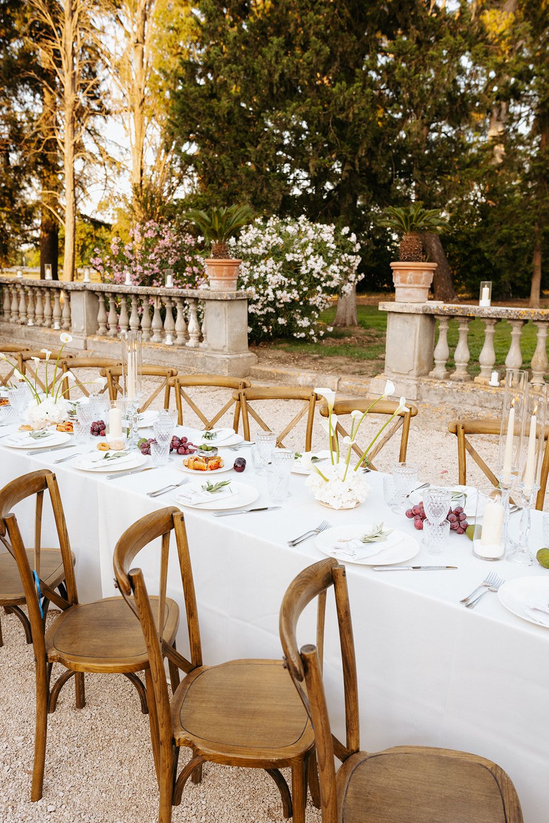 Elegant outdoor dining table with white linens and wooden crossback chairs on a patio with stone balustrades and flowering plants.