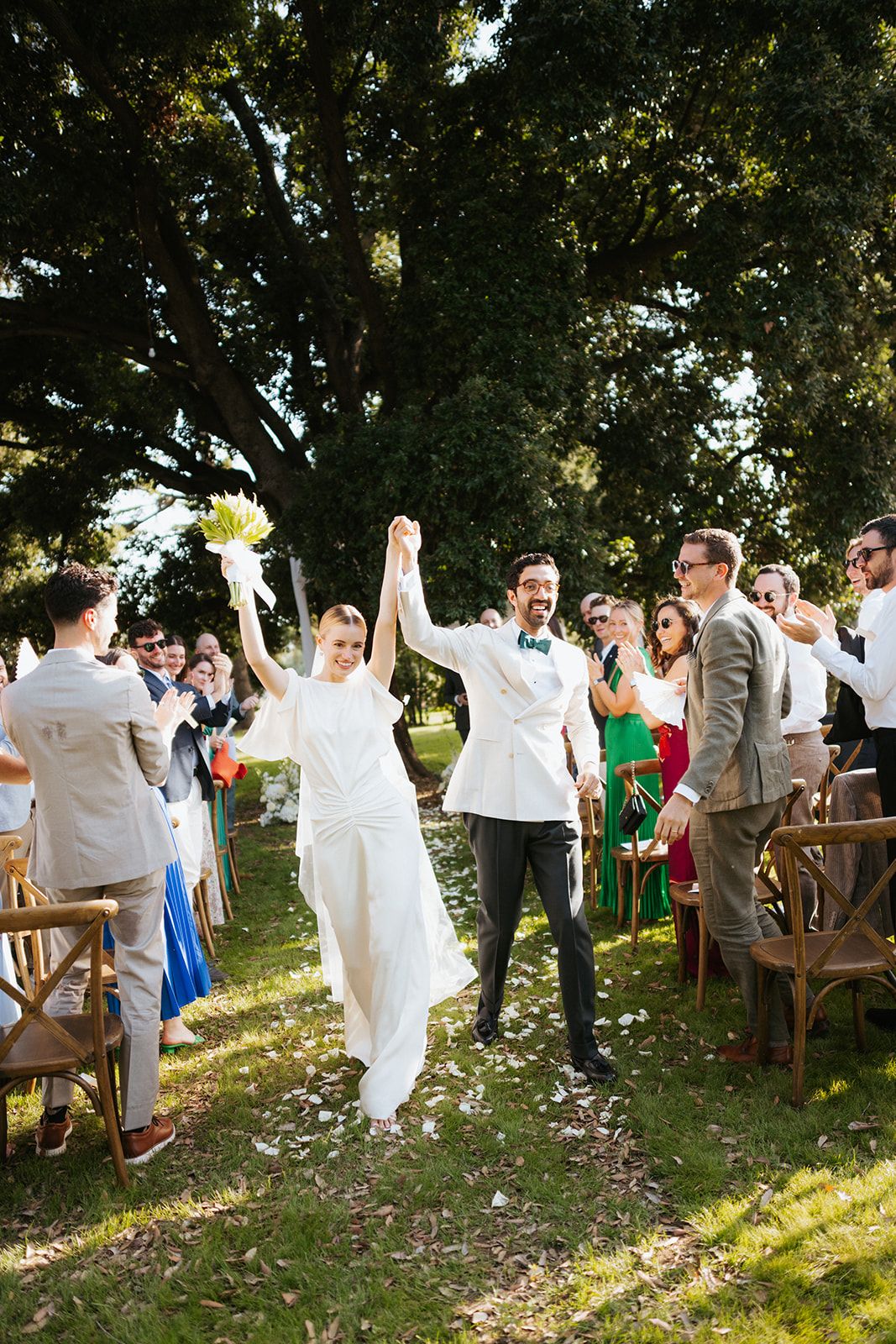 Newlyweds celebrate with guests during outdoor wedding ceremony with flower petals scattering the ground.