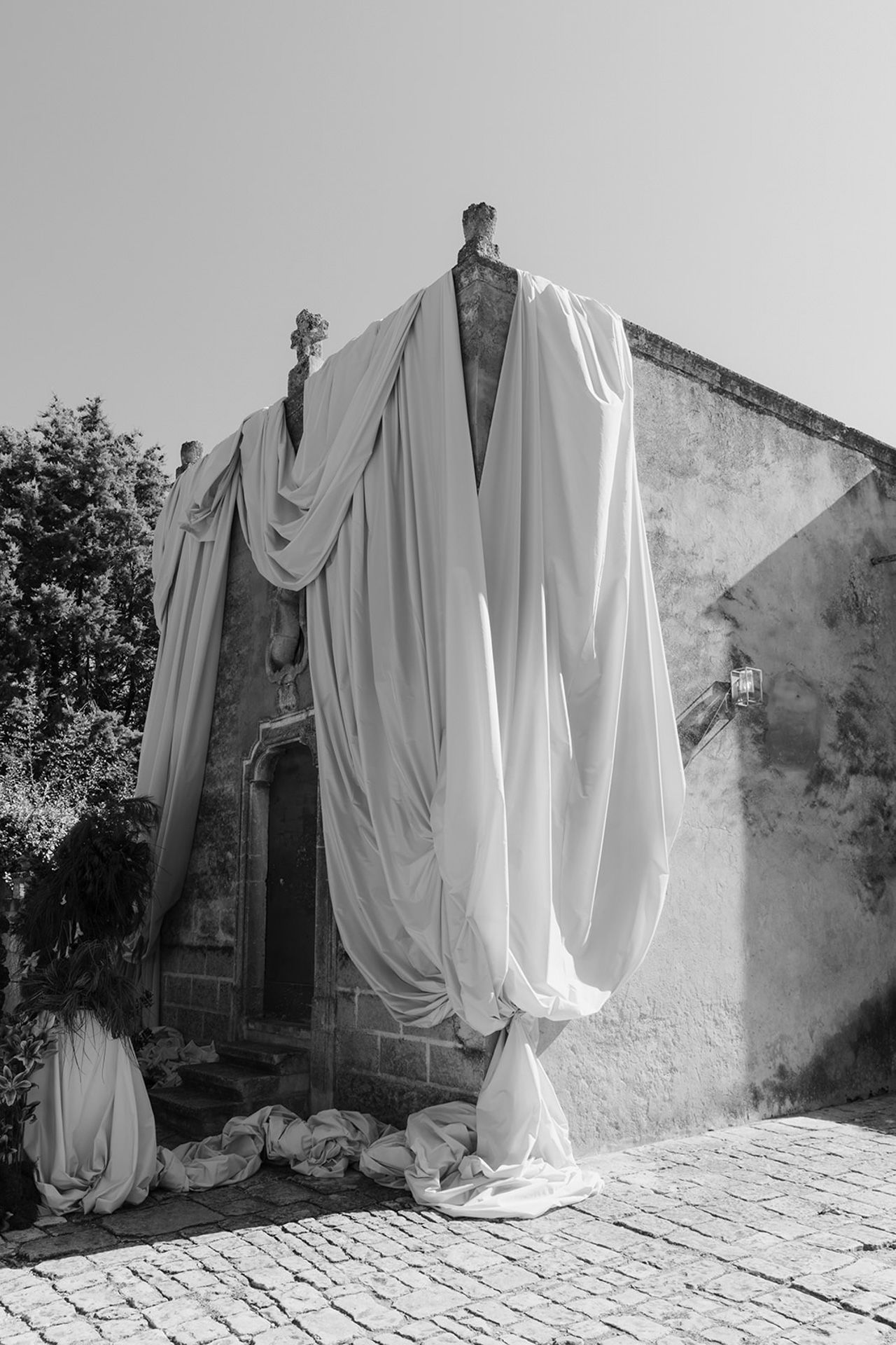 A dramatic black and white photograph of flowing white fabric draped over a mannequin against a weathered stone wall.