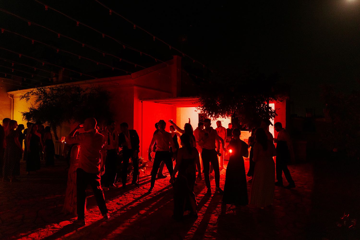 Silhouetted people gather outside buildings at night illuminated by dramatic red lighting creating an eerie atmosphere.