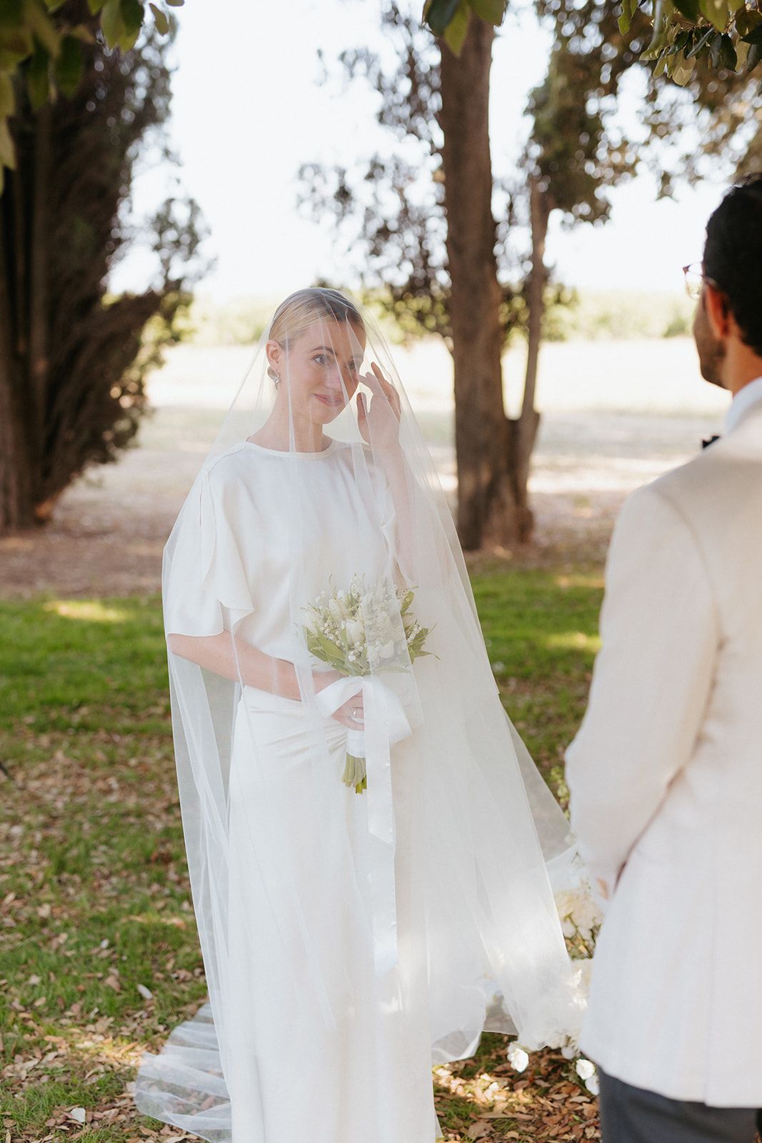Elegant wedding couple stands among trees, with the bride in white dress holding a bouquet during an outdoor ceremony.