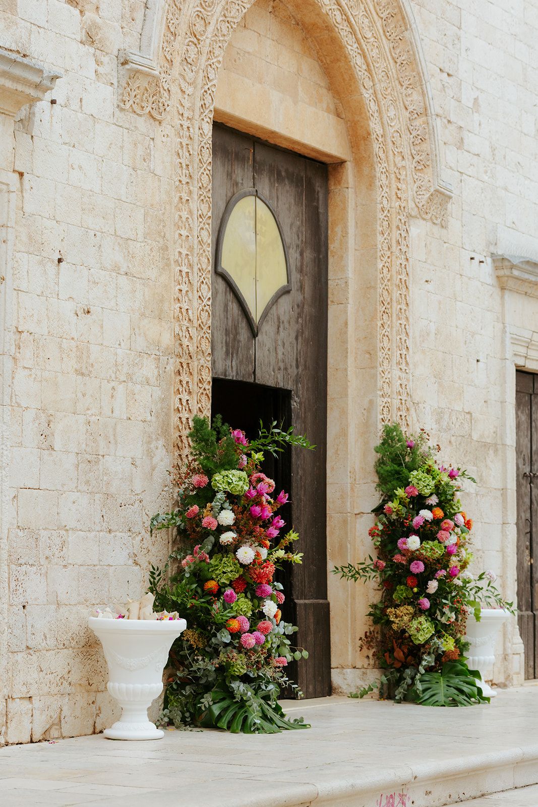 Ornate wooden church doors adorned with lush pink and white floral arrangements against a stone facade.