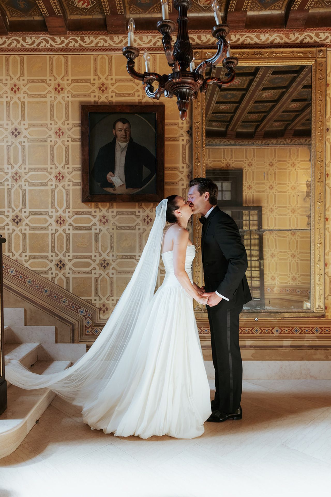 A romantic wedding scene in an ornate historic mansion with patterned walls, chandelier and flowing white bridal gown.