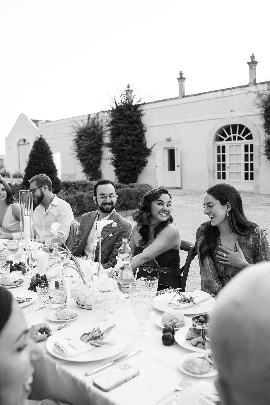 A black and white photo shows wedding guests laughing together during an outdoor reception dinner at an elegant venue.