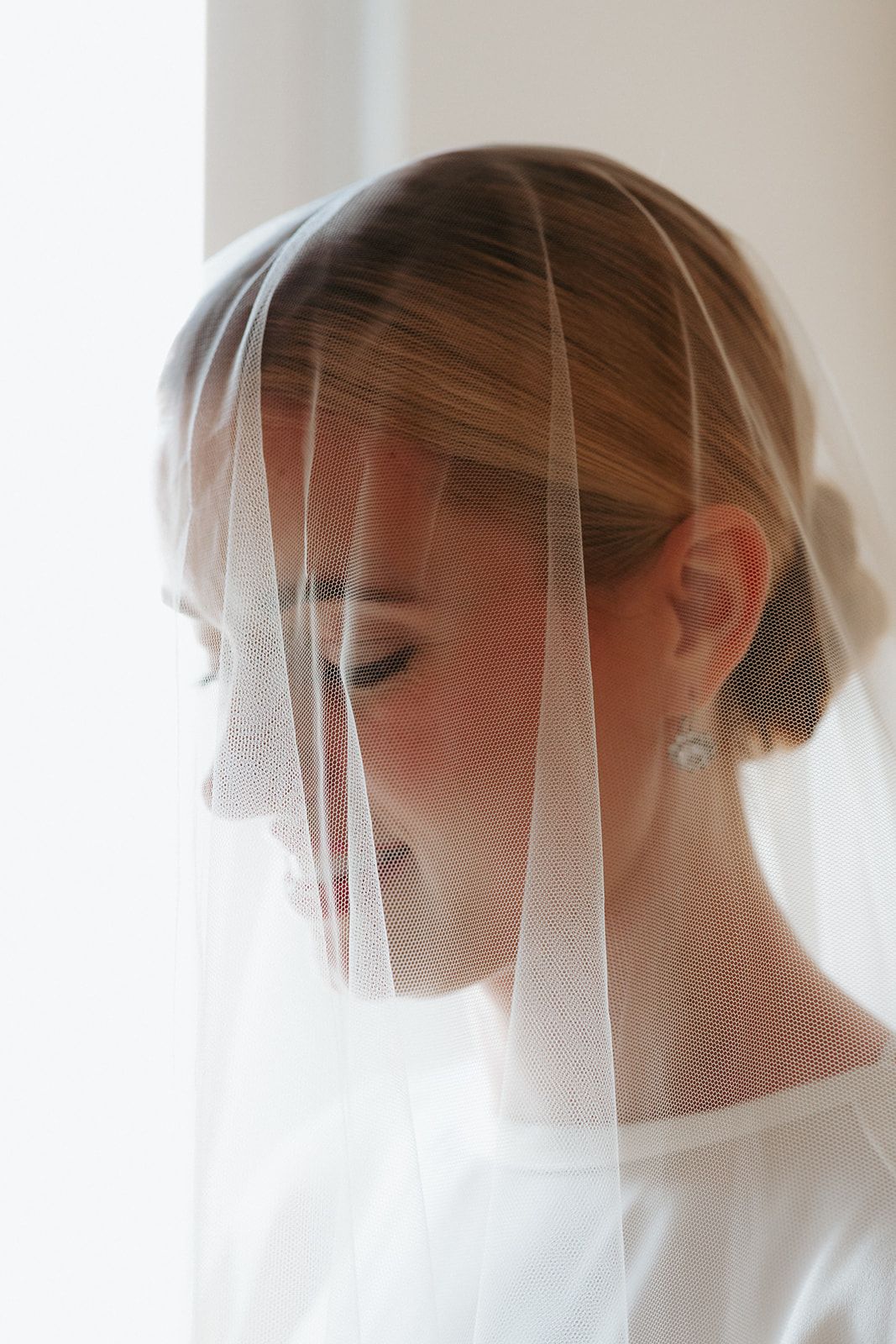 A side profile silhouette of a bride wearing a delicate veil against a bright window background.