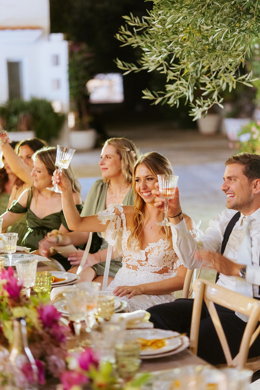 Wedding reception guests seated at a long dining table under string lights and greenery enjoying a festive outdoor dinner.
