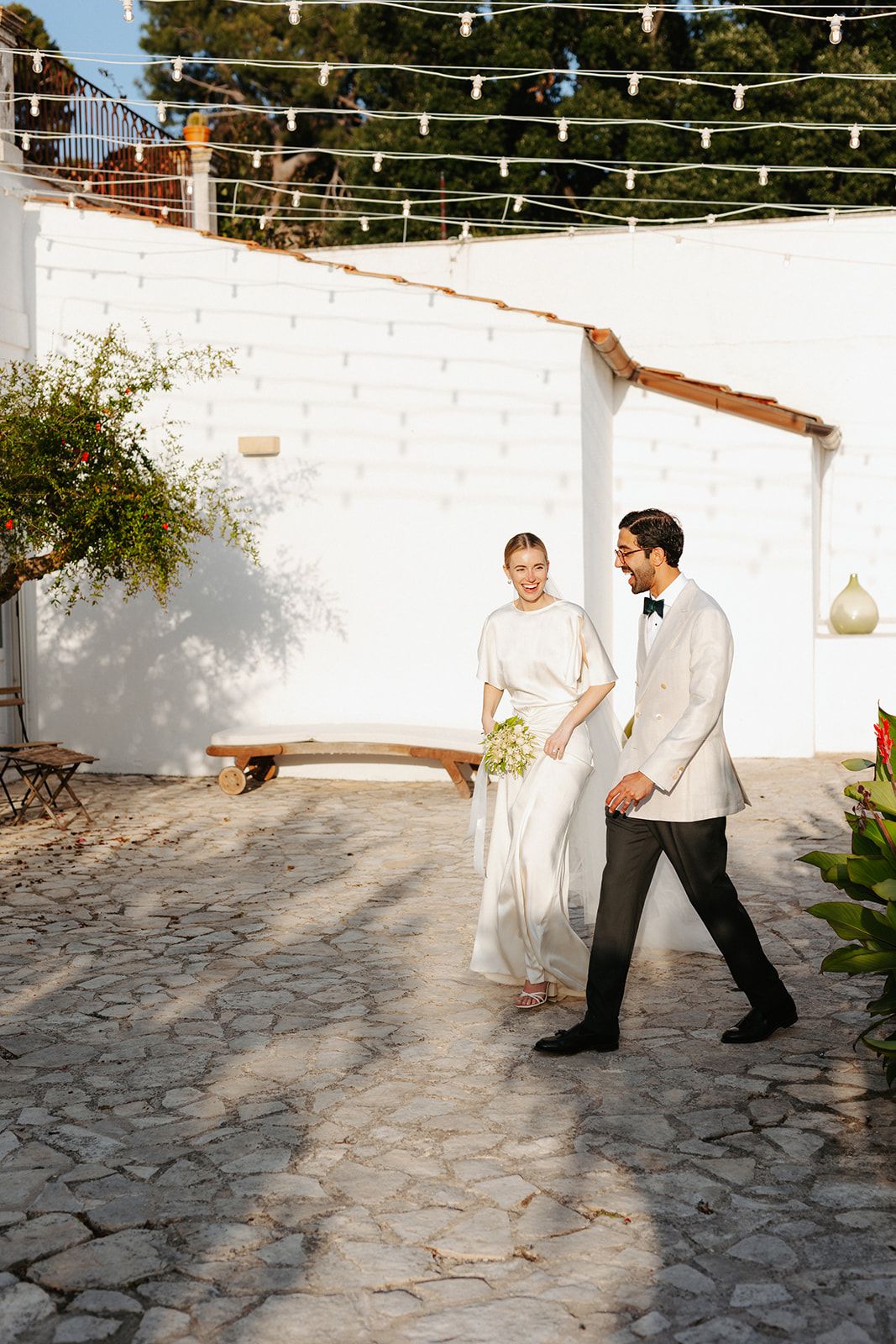 Wedding couple walking on a stone patio with string lights overhead and white stucco walls in Mediterranean setting.