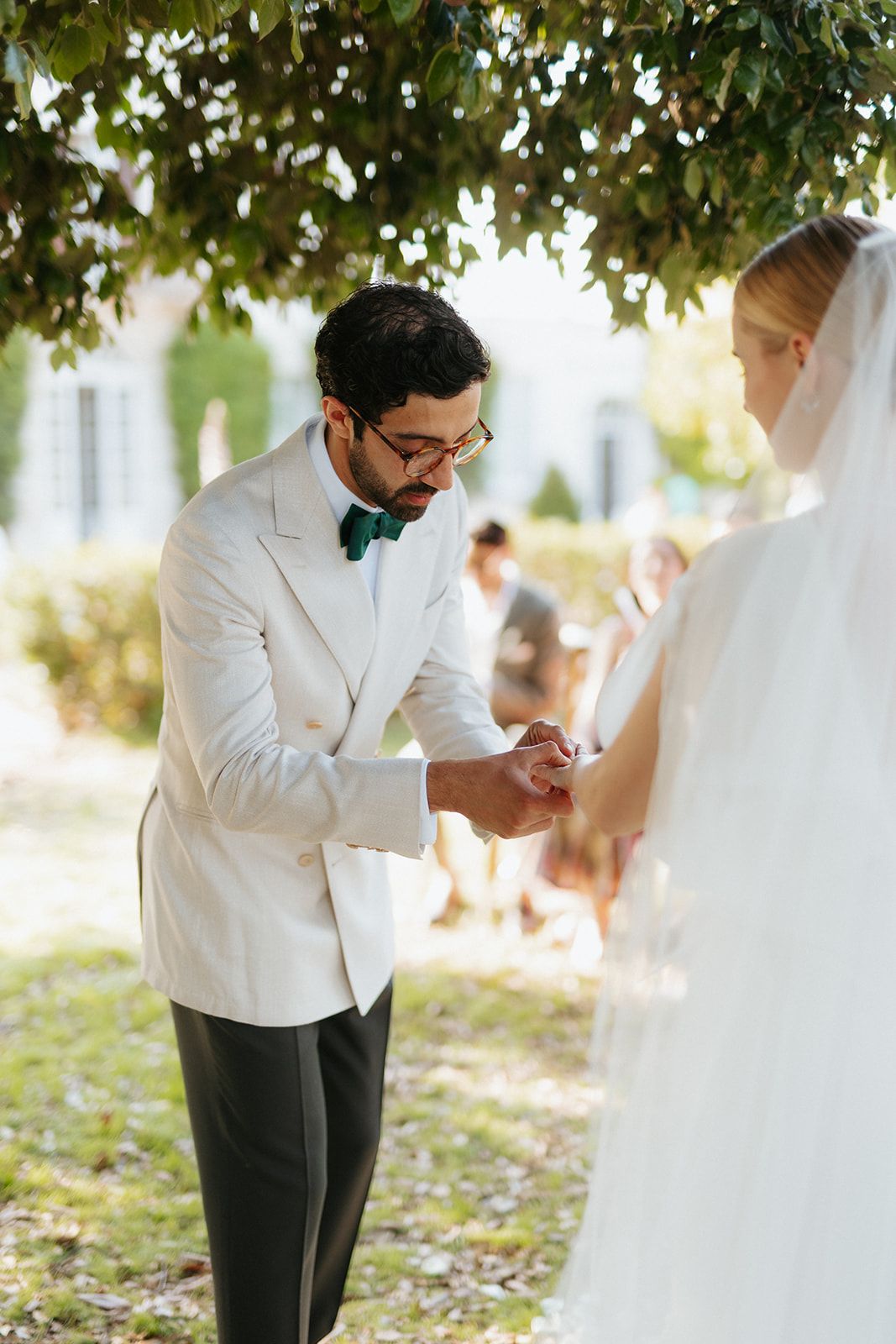Wedding ceremony takes place outdoors under green leaves as groom in white jacket and bow tie exchanges rings with bride.