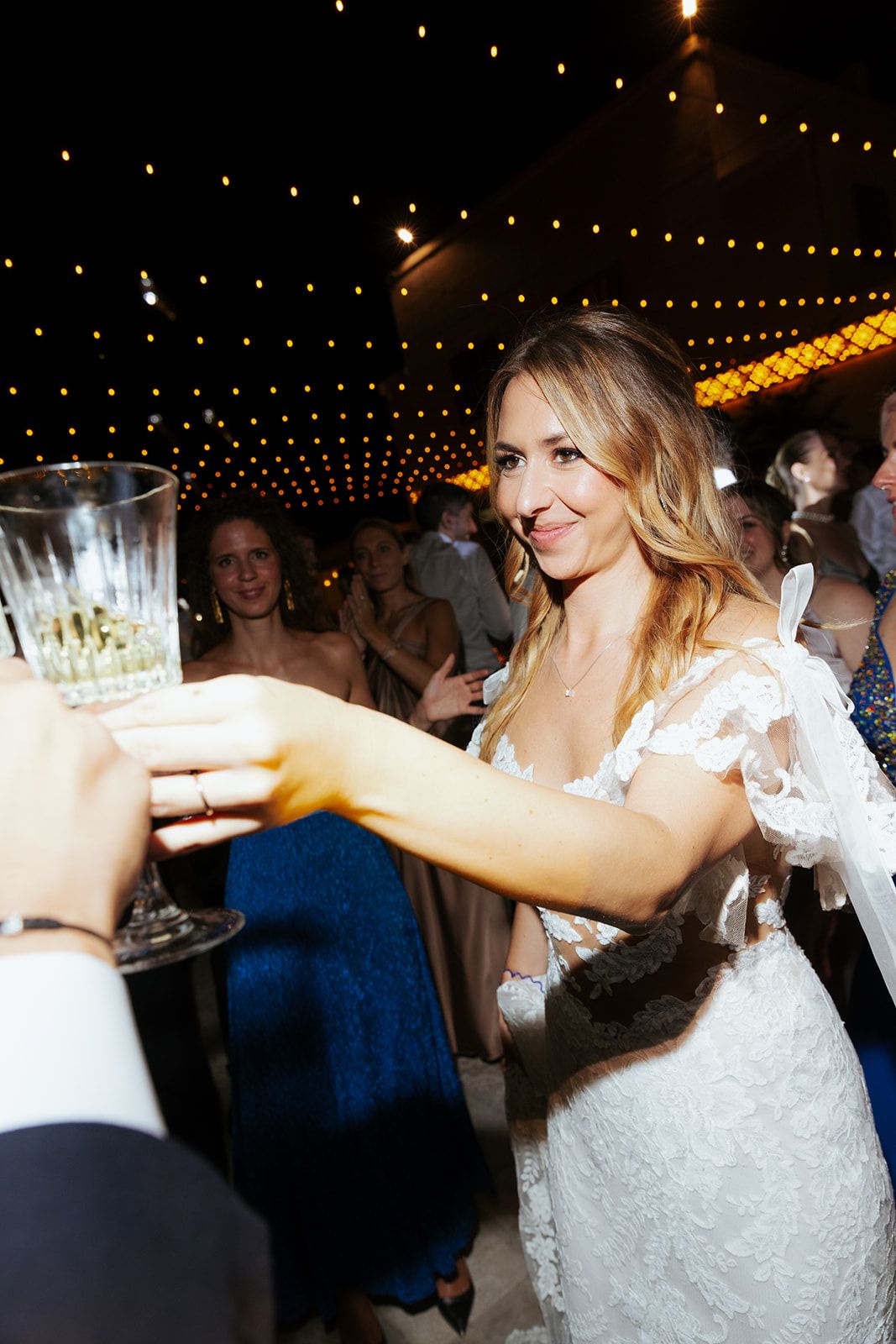A bride in a white lace dress toasts with a champagne glass under twinkling string lights at an evening reception.