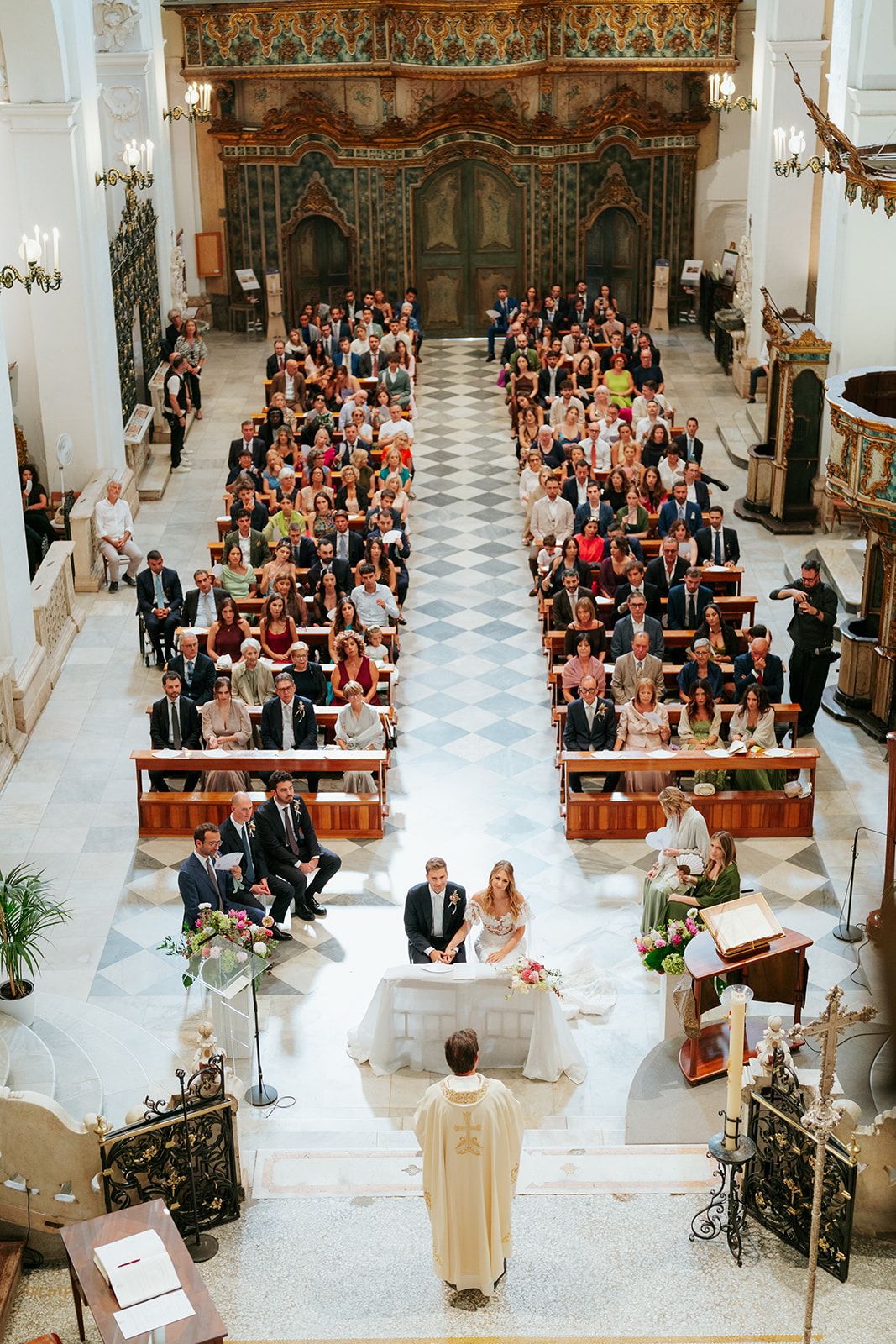 Wide view of a Catholic church wedding ceremony showing guests seated in ornate historic cathedral interior.