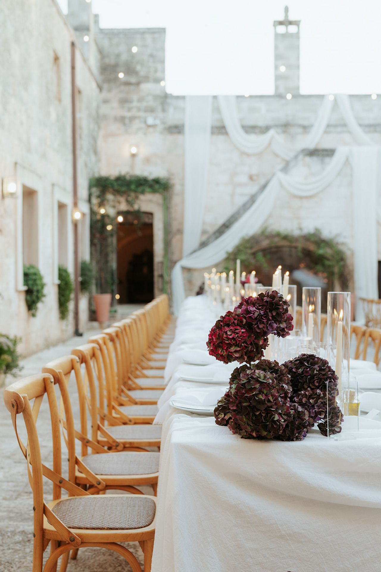 Elegant wedding reception setup with wooden chairs and burgundy floral centerpieces along a white tablecloth in a rustic venue.