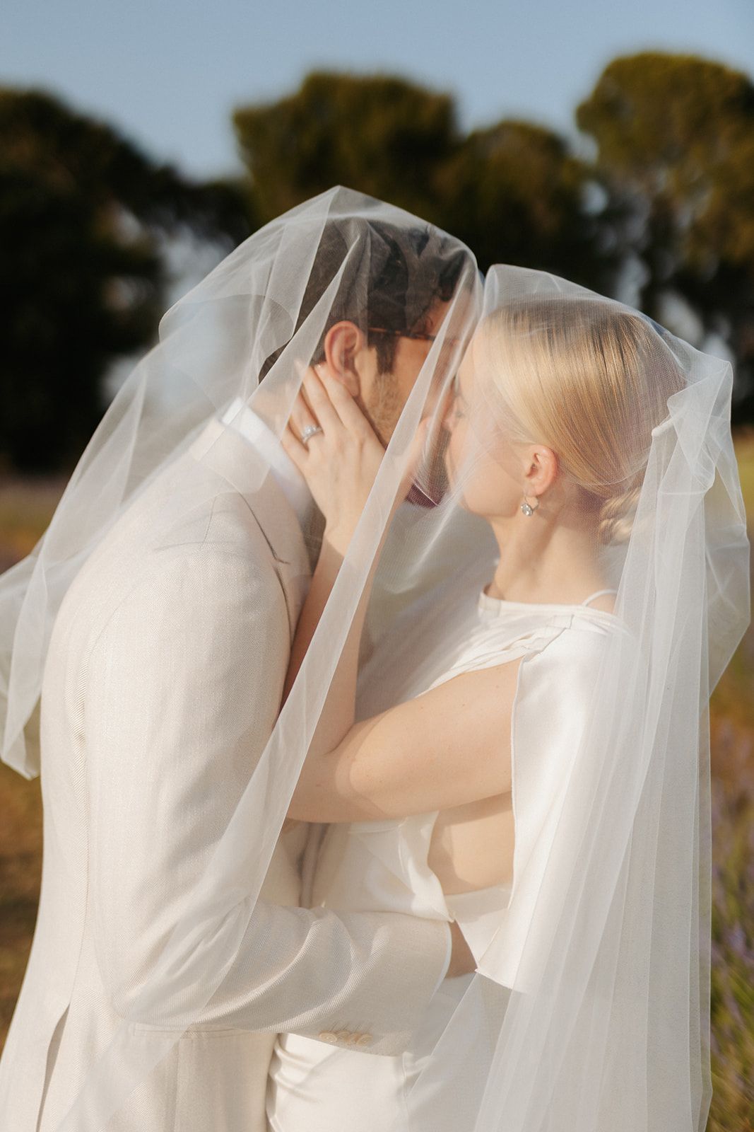 A couple shares an intimate kiss under a flowing white wedding veil in an outdoor garden setting.