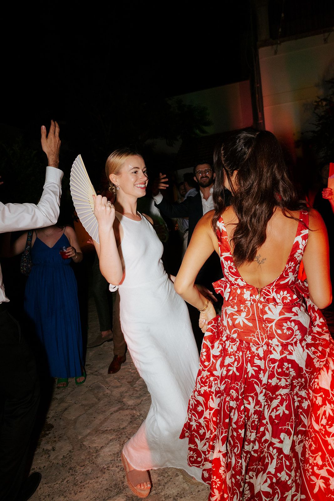 A bride in a sleek white dress dances exuberantly with guests, including someone in a vibrant red floral dress at a reception.