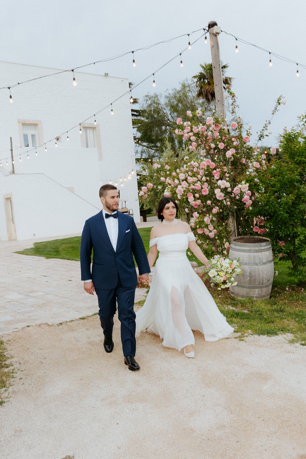 A couple walks hand in hand on a gravel path under string lights with floral decorations at an outdoor wedding venue.