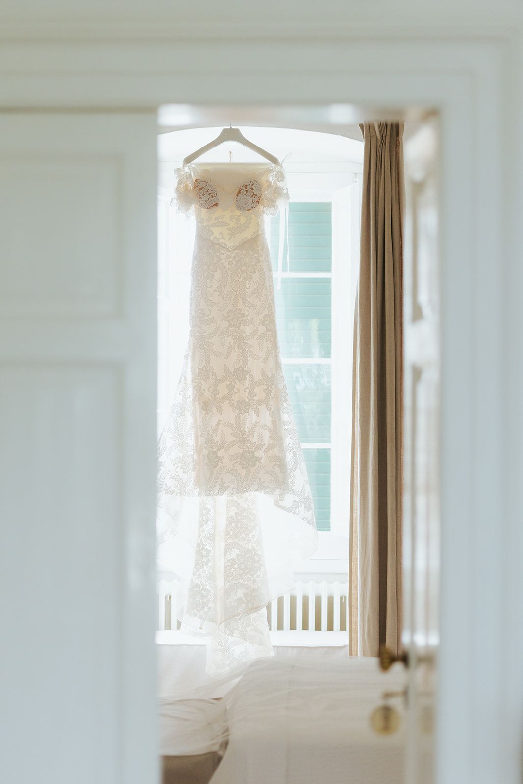 White lace wedding dress hanging by window with soft natural light illuminating delicate details in bright room.