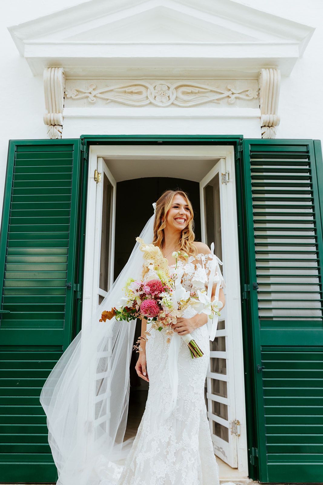 Elegant wedding scene with white window shutters framing someone in a lace gown holding a pink bouquet.