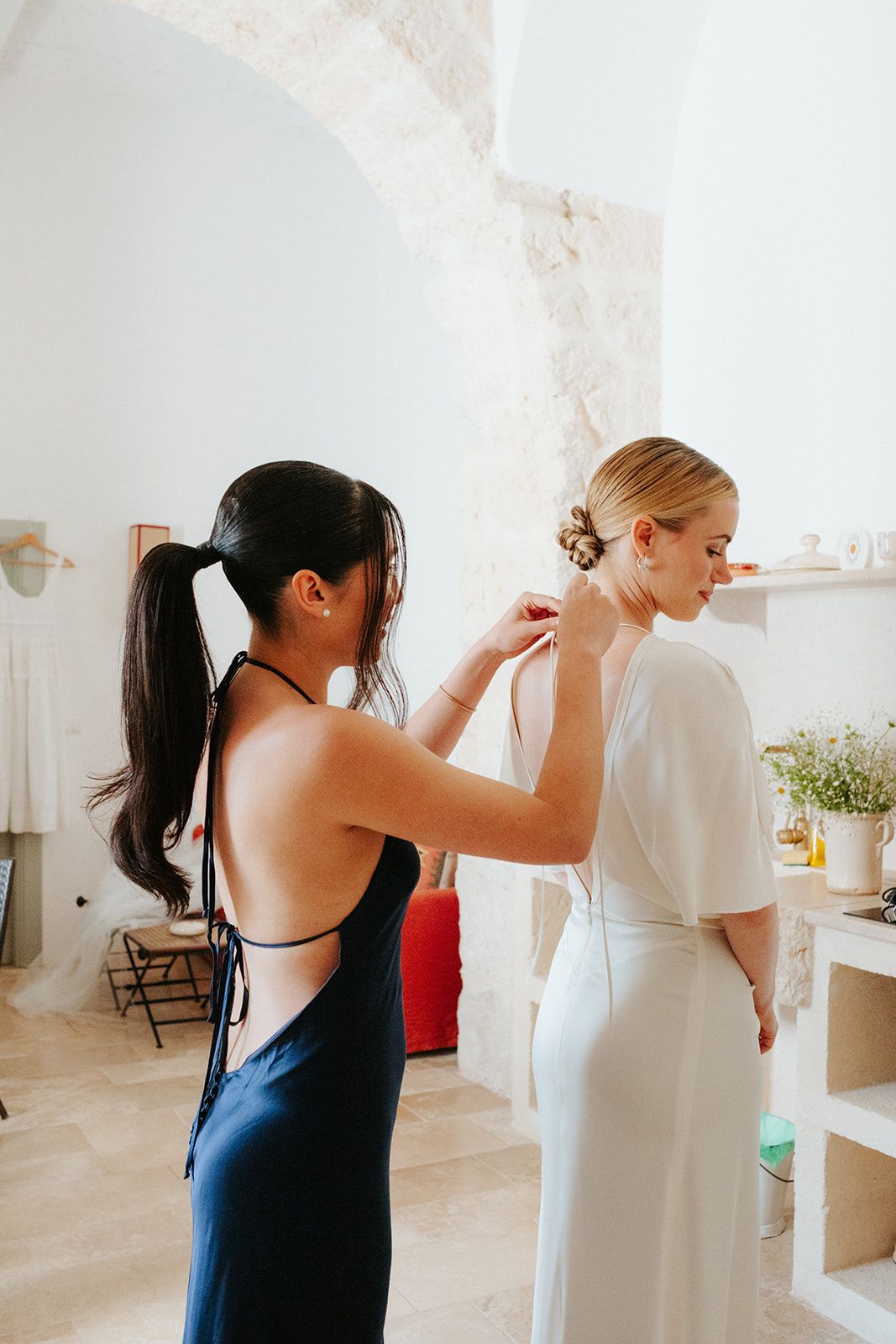 Someone helps with final adjustments to a classic white wedding dress in a bright, sunlit room with minimalist decor.