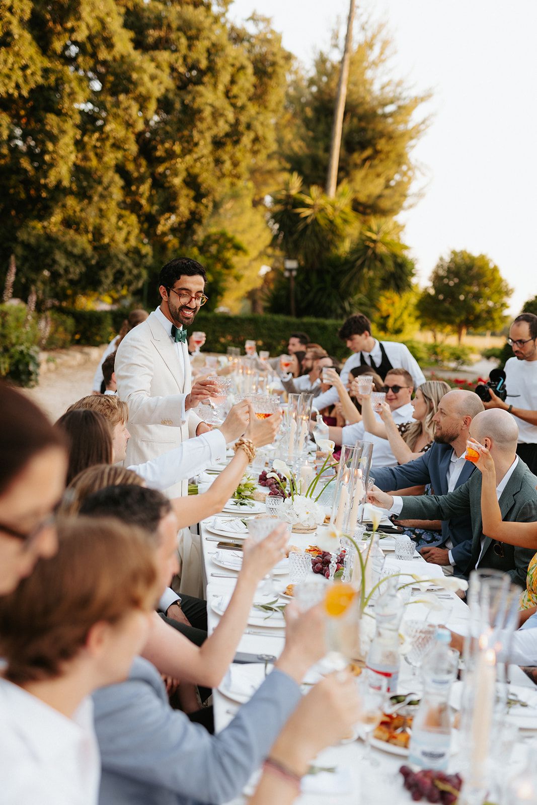 Outdoor wedding reception dinner with guests raising glasses in a toast at long elegant table during golden hour.