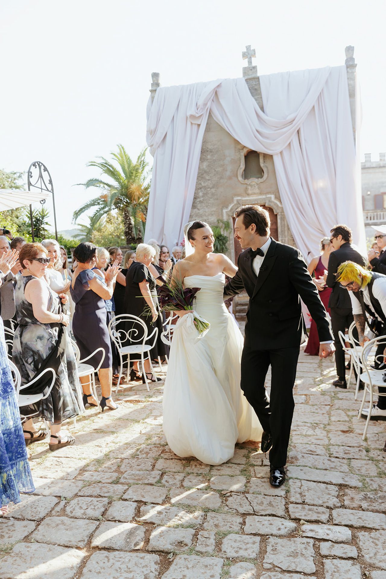 Wedding guests dancing and celebrating on a stone patio with white drapes and palm trees in the background.