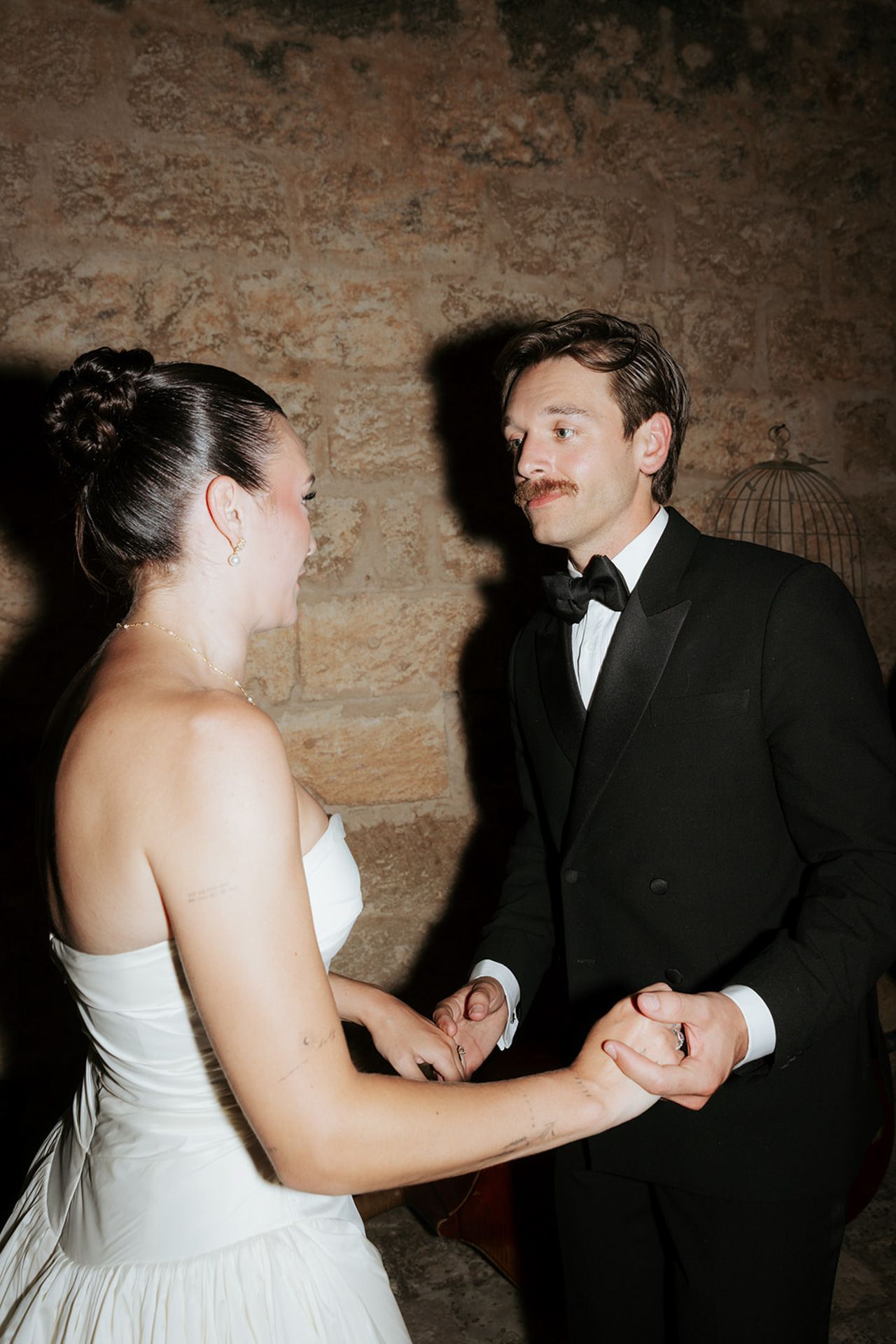 A bride and groom dance together in a dimly lit room with rustic stone walls in the background.