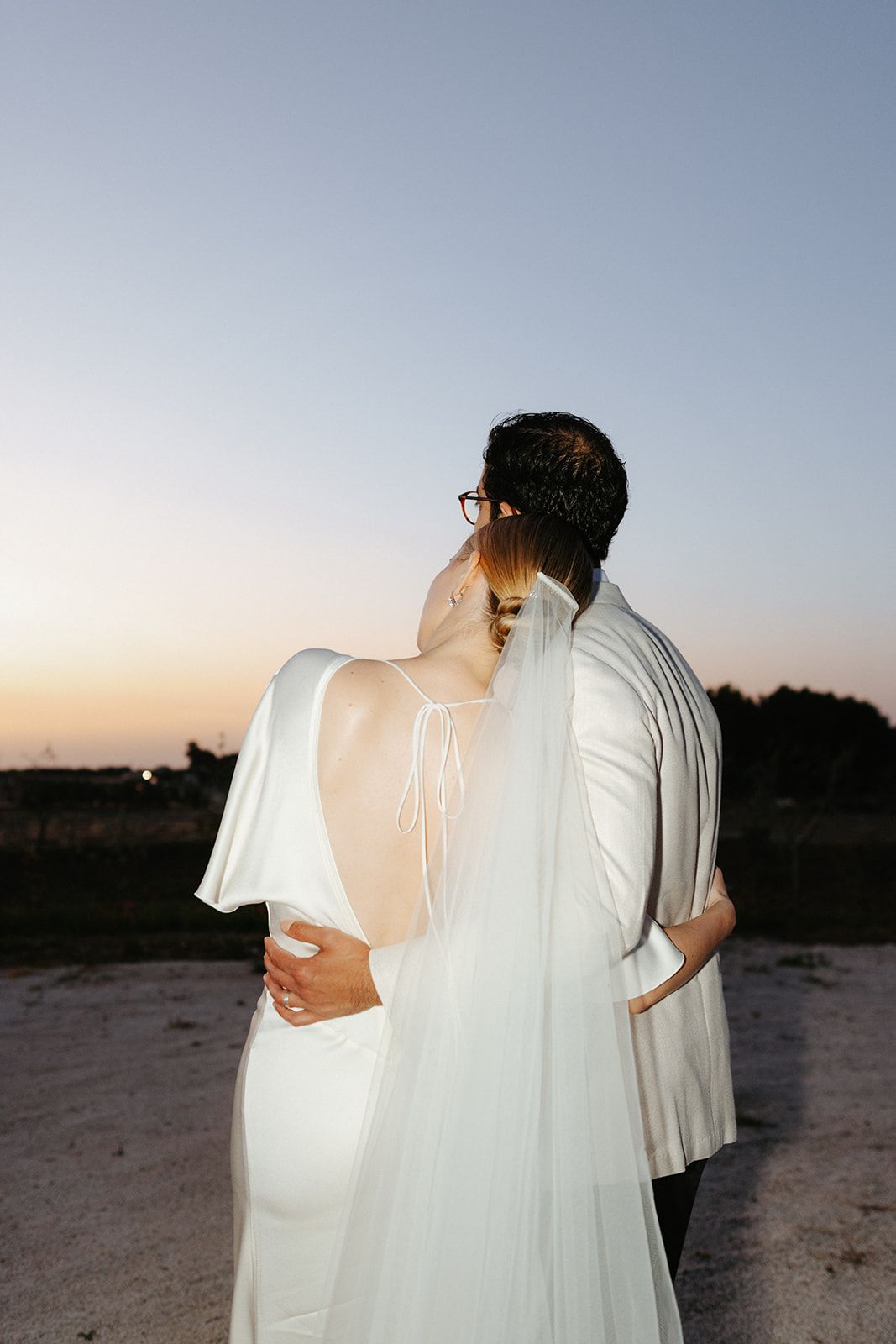 A couple in white formal attire embraces while watching the sunset on a dirt path during golden hour.
