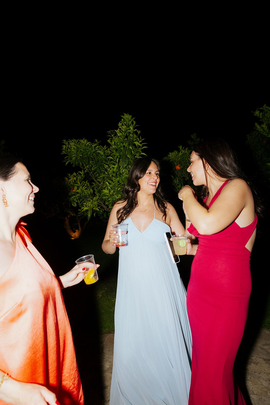 Three people in colorful formal attire laugh and chat while holding drinks at an outdoor nighttime event.