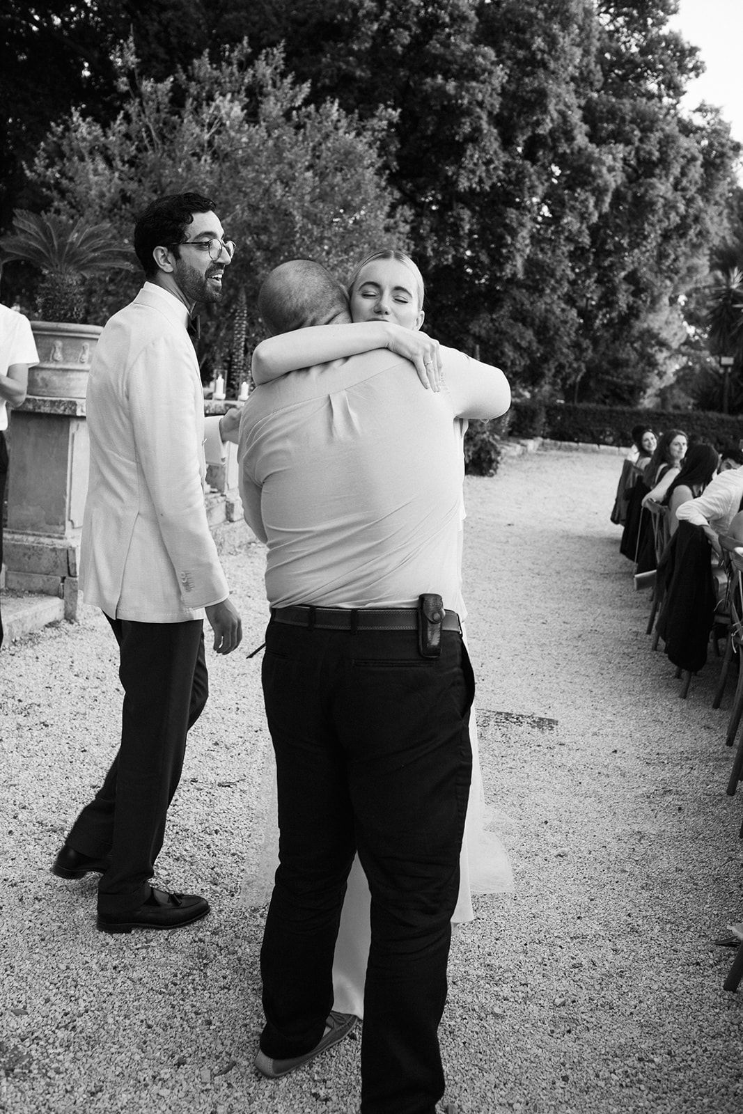 Black and white candid photo of people hugging and celebrating at an outdoor event on a gravel path.