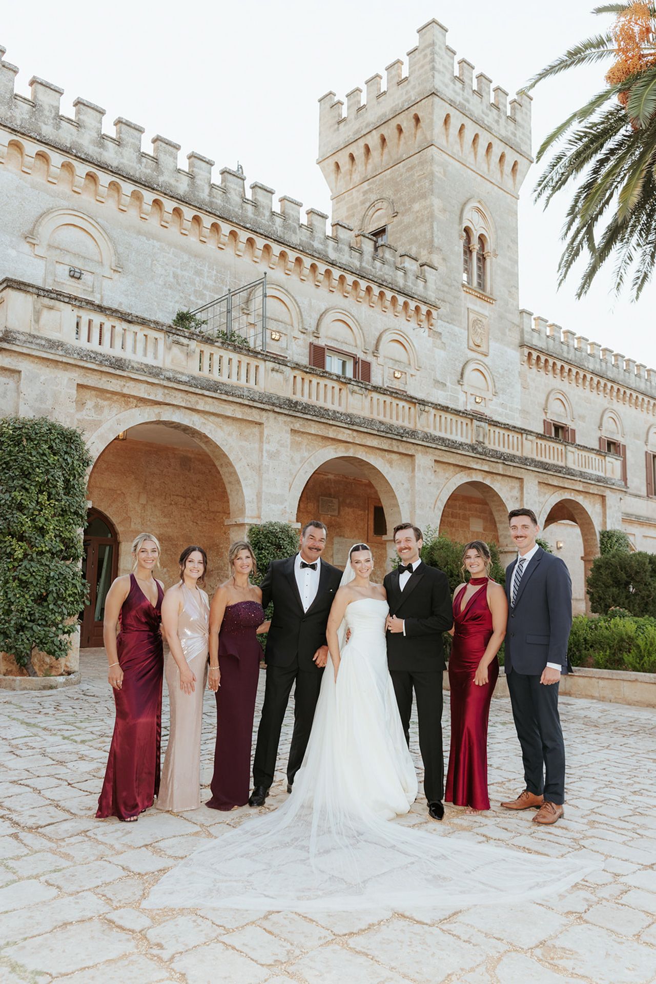 Elegant wedding party poses in front of a majestic Mediterranean castle with stone archways and palm trees.
