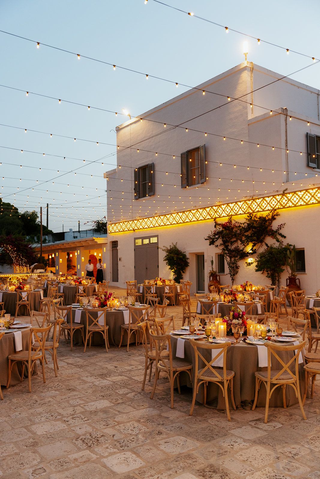 Elegant outdoor reception venue with string lights, wooden chairs and tables arranged on a stone patio at dusk.