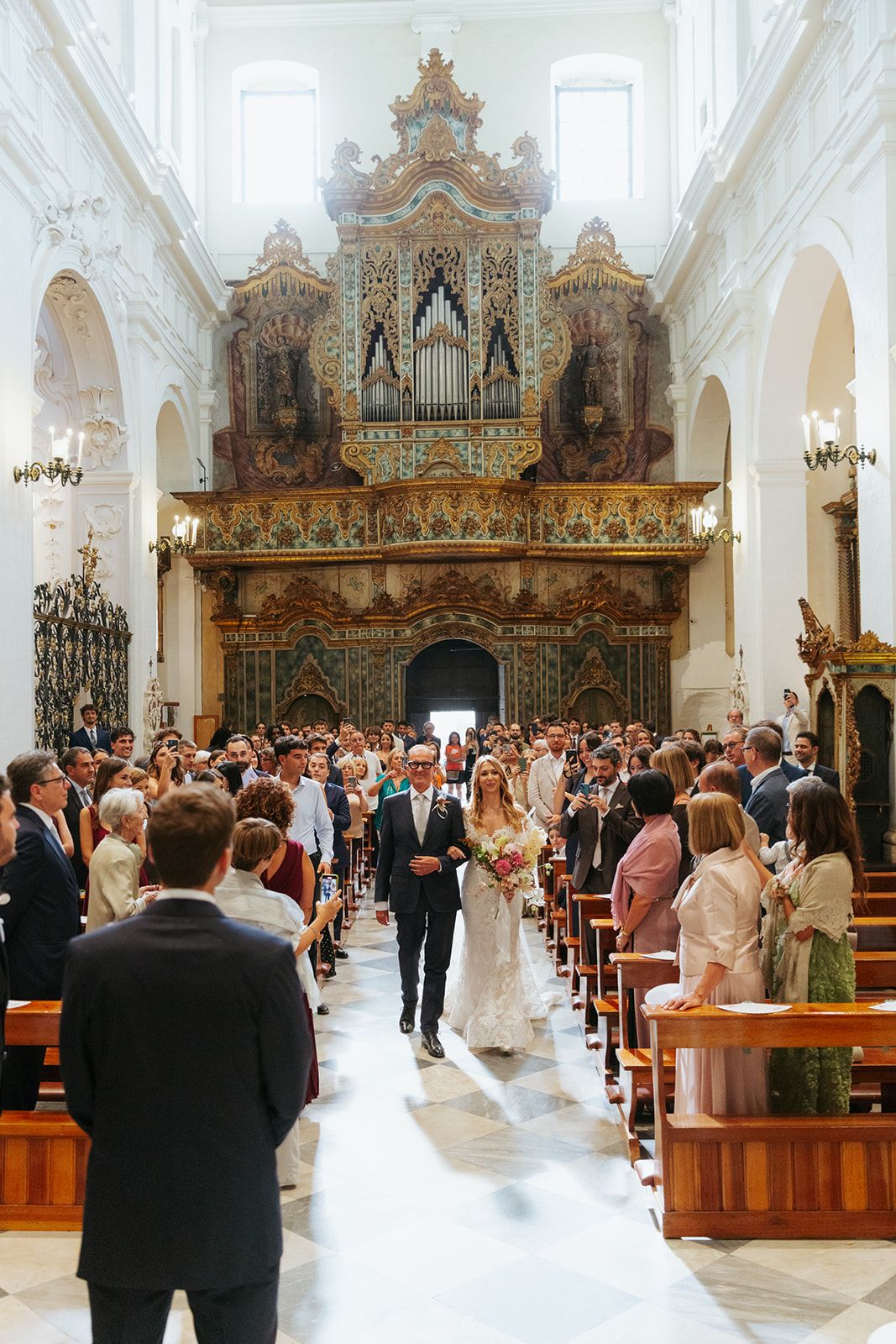 Interior view of grand Catholic church wedding ceremony with ornate baroque organ and wooden pews filled with guests.