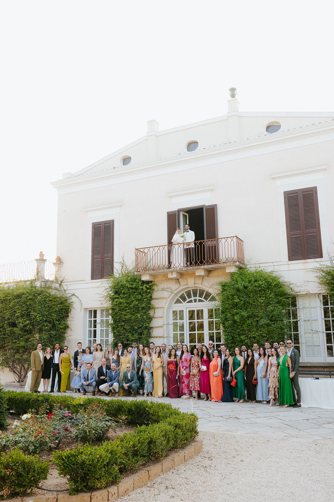 Large group of wedding guests dressed in vibrant attire gather in front of a white Mediterranean-style villa.
